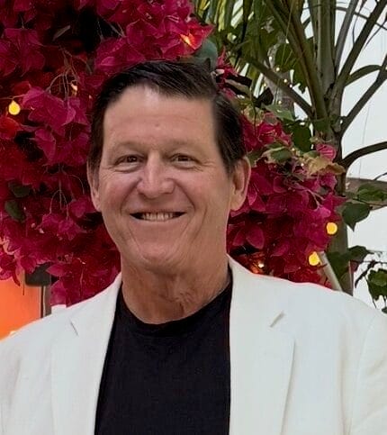 Headshot of Ben Starrett, a man with white skin and brown hair wearing a white jacket and black shirt, standing in front of flowers.