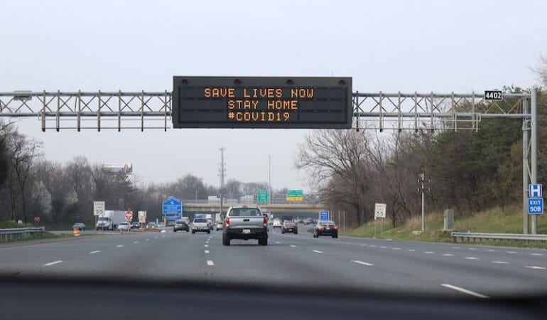 A sign on a highway reads "Stay Home" as cars drive underneath it.