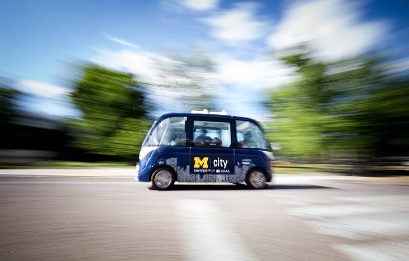 This image shows a street with a blue driverless shuttle in the middle. Blurred trees sit in the background of the image.