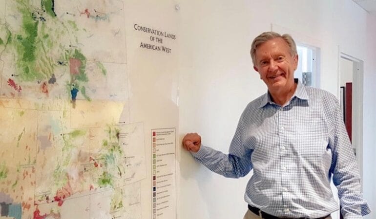 A man in a dress shirt stants to the side of a map showing the topography and geography of the Western United States.