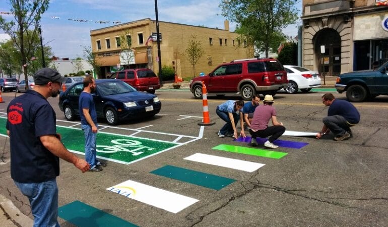 This picture shows several people laying down a new colorful crosswalk on a paved road.