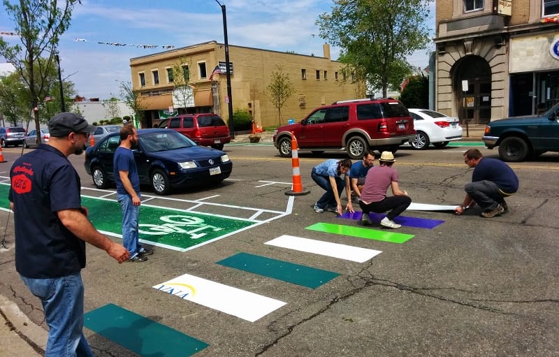 This picture shows several people laying down a new colorful crosswalk on a paved road.