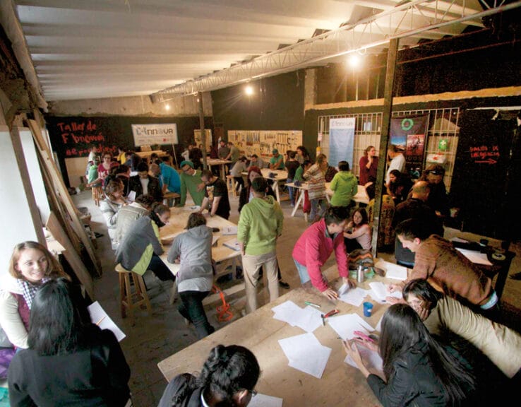 Photo shows the interior of a open room -- C-Innova's makerspace in Bogotá -- with several work tables and at least 30 people standing