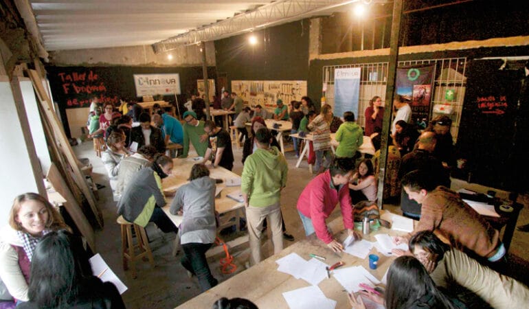 Photo shows the interior of a open room -- C-Innova's makerspace in Bogotá -- with several work tables and at least 30 people standing