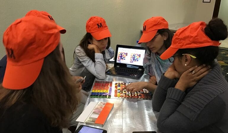 A group of participants in a Lincoln Institute course wear orange hats and gather around a game board. The hats say "M" for "middle class."