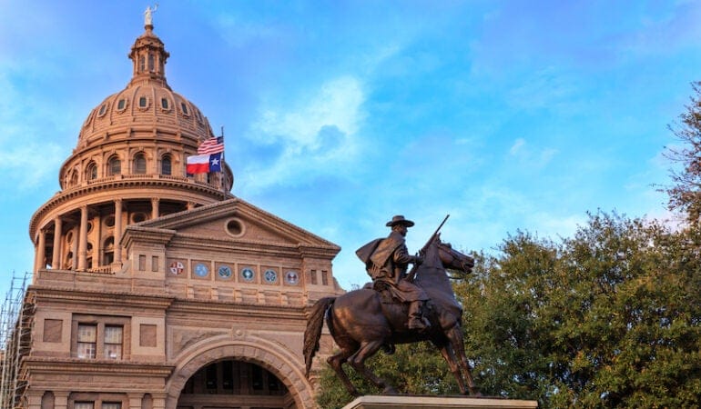 A sand-colored building with a domed roof stands on the left with trees on the right and a statue of a man on a horse in the foreground.