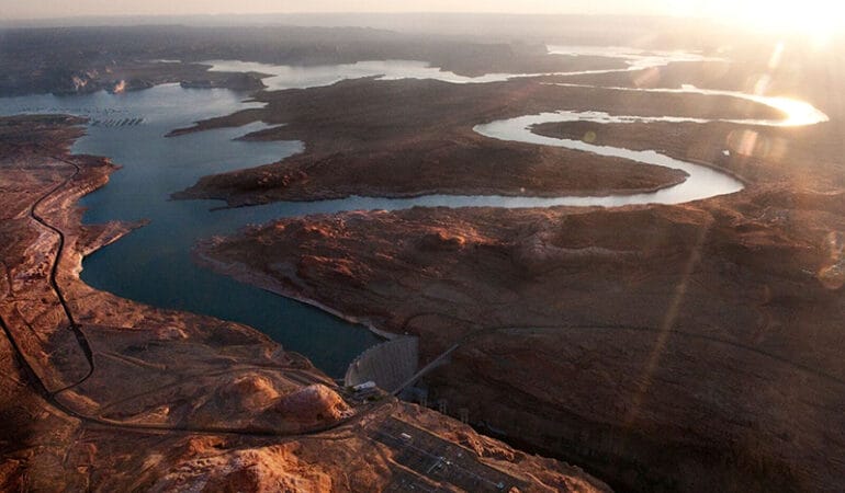 The sun glares white in the top right corner over a landscape of river and desert viewed from above. Two main branches of the Colorado river appear silver as they wind across the land and reflect the sun. They merge with the irregular