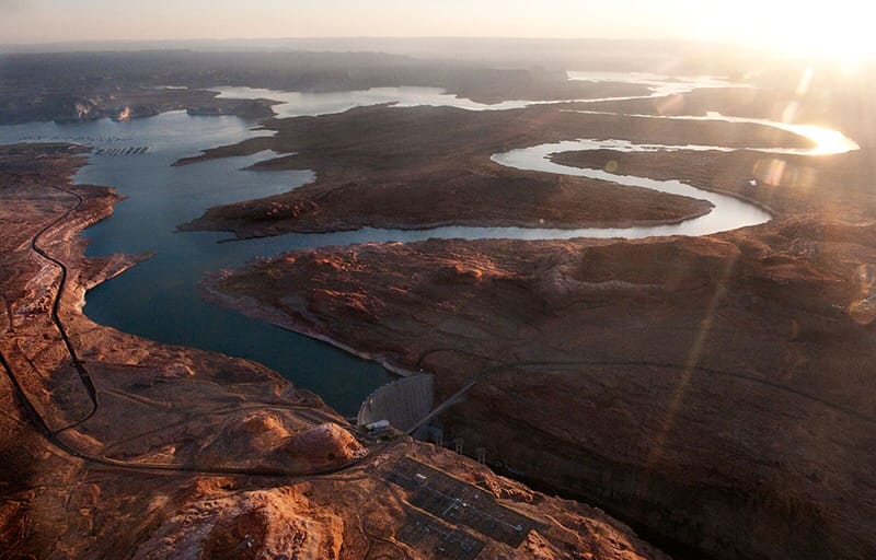 The sun glares white in the top right corner over a landscape of river and desert viewed from above. Two main branches of the Colorado river appear silver as they wind across the land and reflect the sun. They merge with the irregular