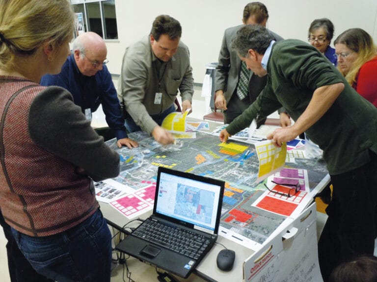 Photograph of several people standing around a table on which there is a colorful plan showing streets and buildings. Some of the people lean toward the surface of the table to place colored stickers on the plan. A laptop computer on the table shows another version of the plan.