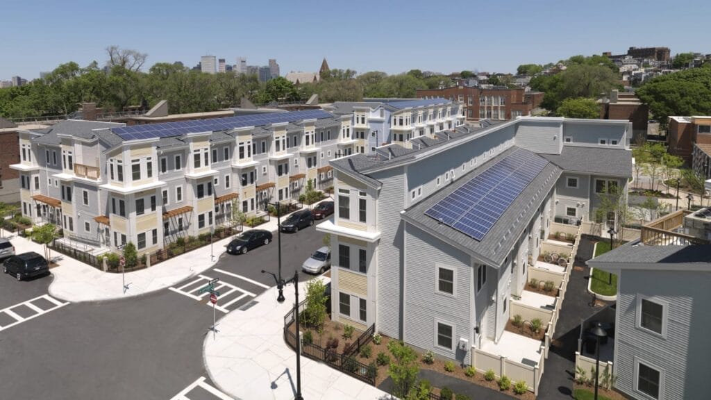 Two rows of tan and gray townhomes with rooftop solar panels. The rows are separated by a paved road flanked by wide,, white sidewalks.