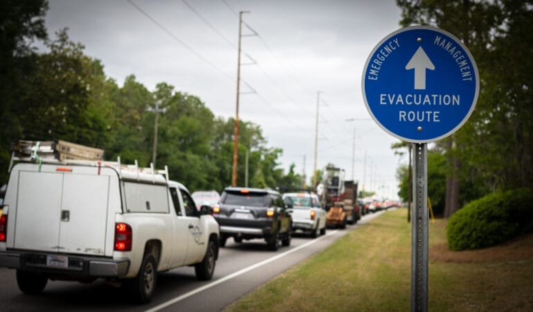 A line of cars drives past a blue sign that reads evacuation route.