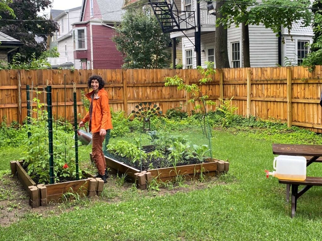 A woman in an orange jacket waters plants in a garden.