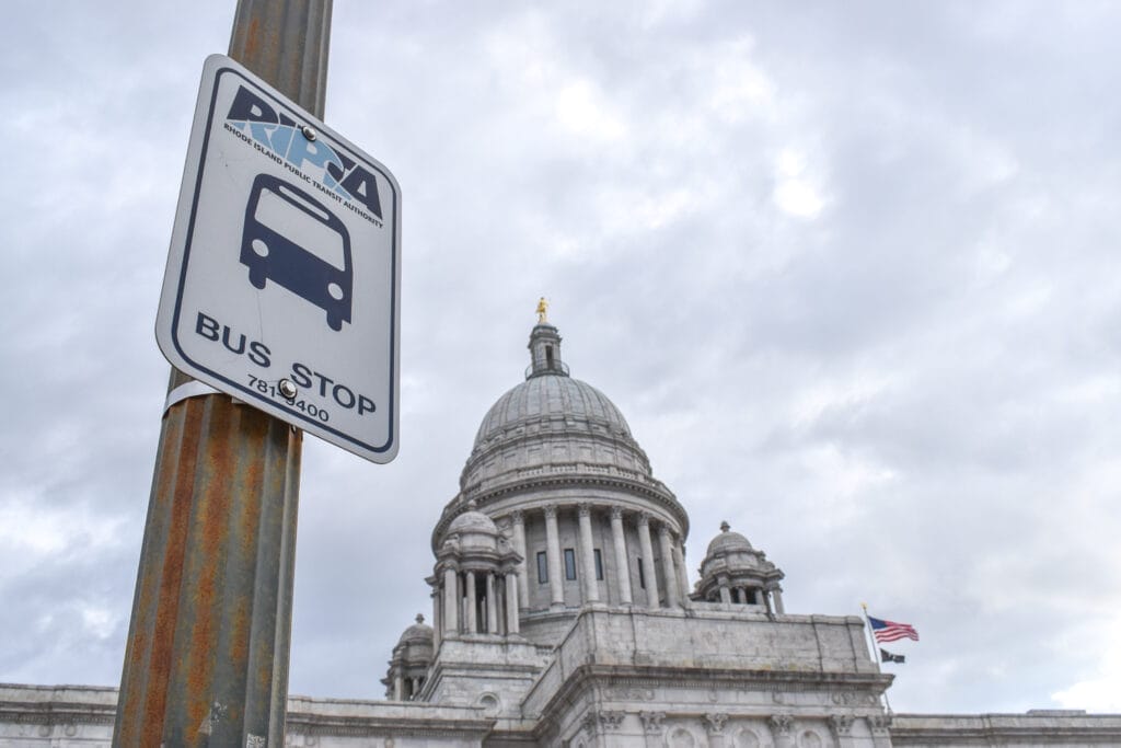 A Rhode Island Public Transit Authority sign on a pole in the foreground with the Rhode Island State House in the background, against a cloudy gray sky.