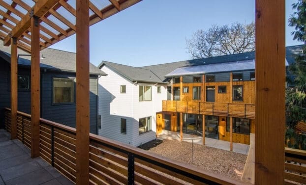 A wooden railing with horizontal slats in the foreground frames a courtyard, a distant building, and blue sky. One section of the building has white siding, and one section has wooden siding.