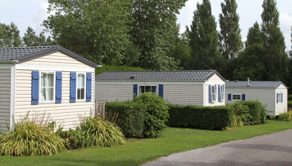 A row of manufactured homes surrounded by grass and green shrubs with trees in the background against a blue sky.