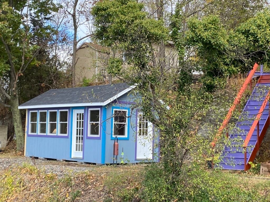 Una casa diminuta azul rodeada de árboles. Detrás de la casa diminuta, se ven casas más grandes.
