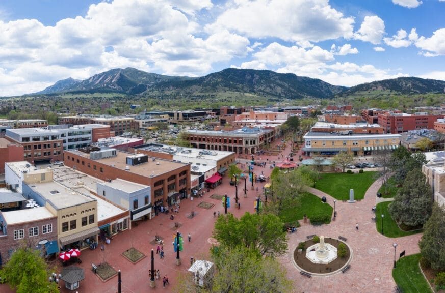 A downtown, pedestrian-only street with lots of shops and restaurants. Large, tree-dotted mountains are in the foreground.
