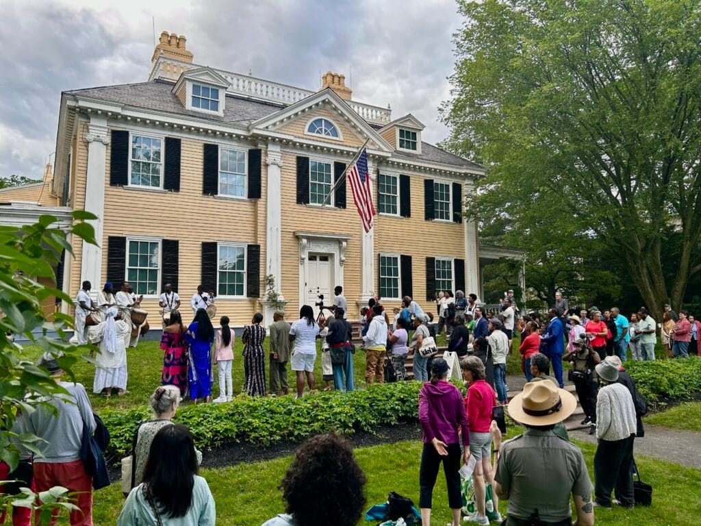 A photo of a crowd of people watching drummers perform at a Juneteenth celebration at the Longfellow House. The picture is taken from behind the crowd, which includes a National Park Service ranger in uniform. A US flag is flying above the front door of the house.