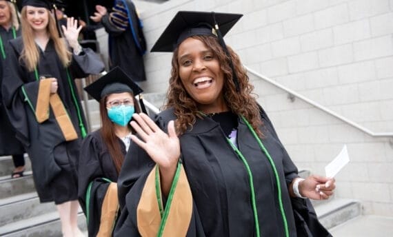 Photo of woman participating in Claremont Lincoln University Commencement wearing a cap and gown and waving.