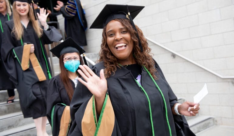 Photo of woman participating in Claremont Lincoln University Commencement wearing a cap and gown and waving.
