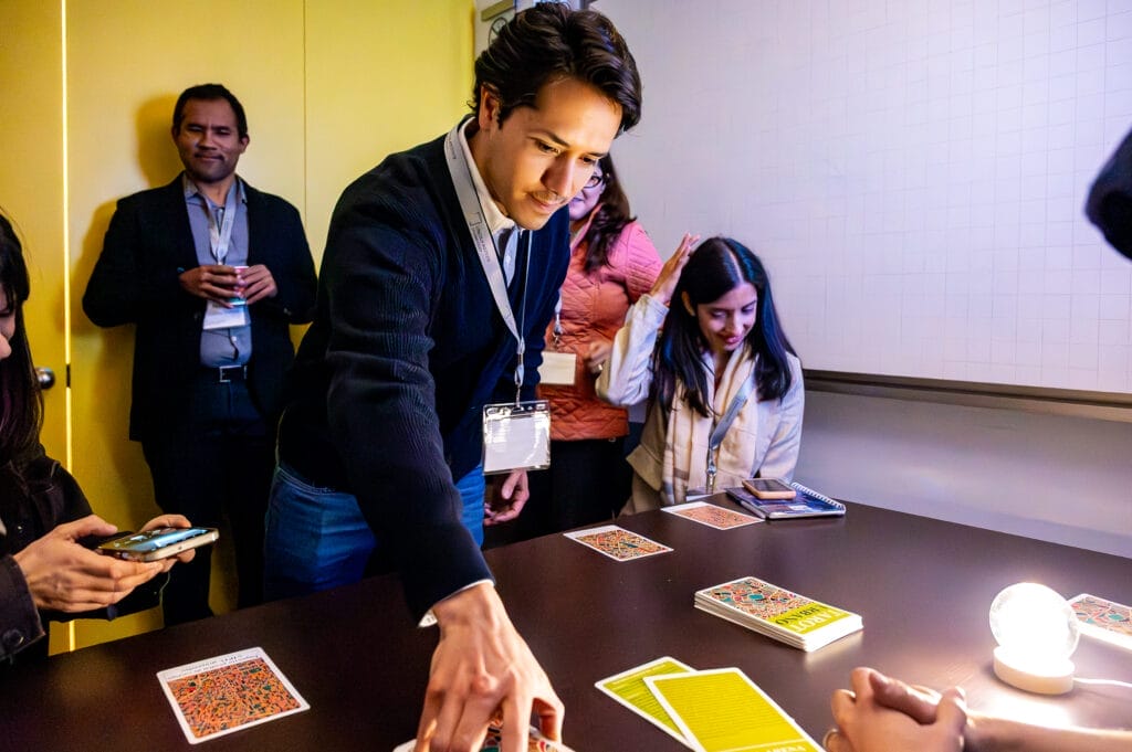 Un hombre elige cartas del Tarot Urbano mientras sus compañeros lo observan. Las cartas están colocadas boca abajo sobre una mesa. Una bola de cristal brilla a lado de ellas.