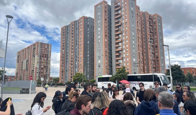 Un grupo de participantes realiza una visita de campo. El grupo acaba de bajarse del autobús y se está reuniendo antes de empezar su visita del plan parcial El Ensueño. En el fondo, se ven los edificios altos que el grupo va a visitar.