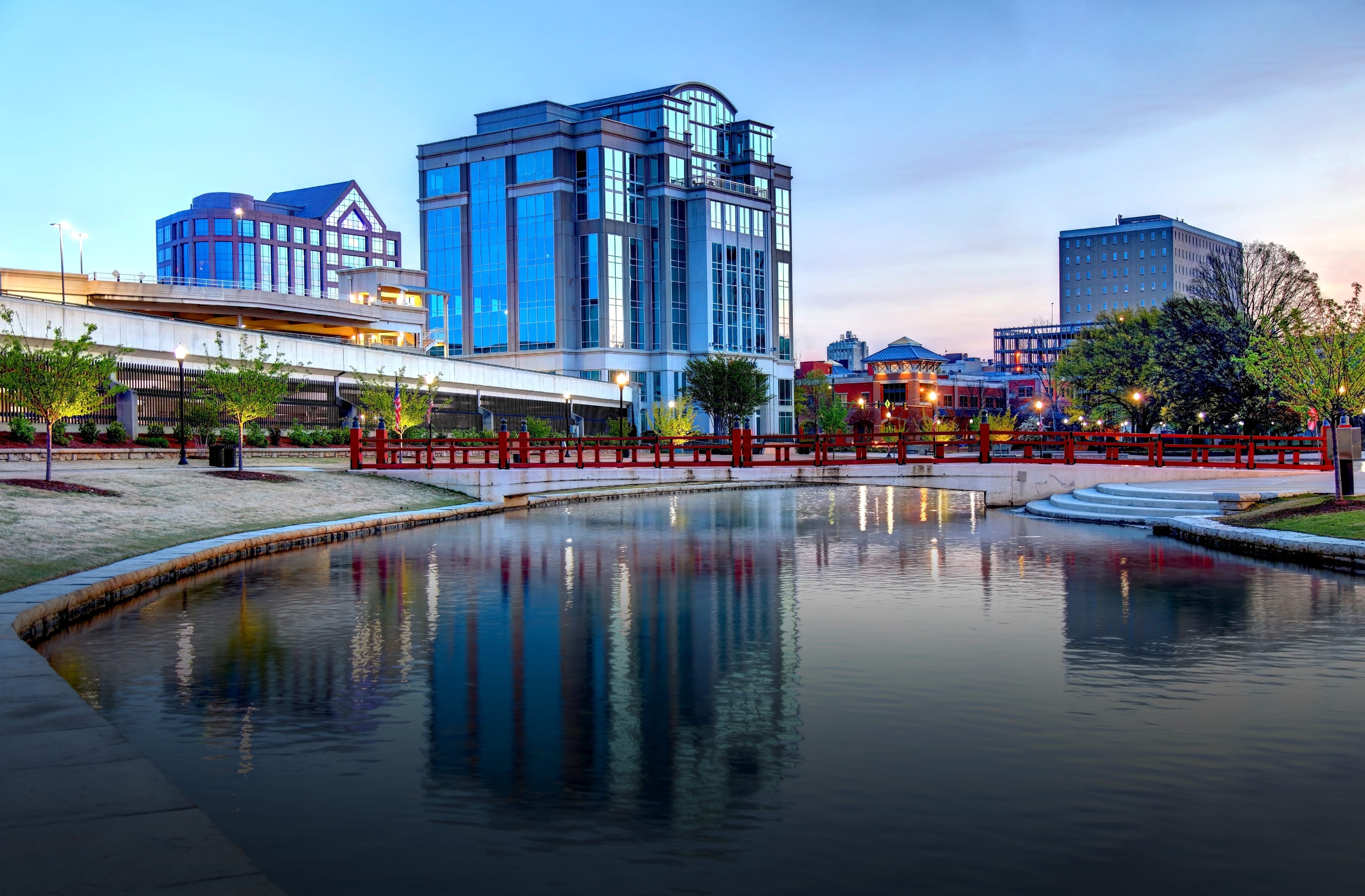 The serene Huntsville, Alabama, cityscape at dusk featuring a reflective canal bordered by a curved stone walkway and a red pedestrian bridge. Modern office buildings with glass facades and traditional brick buildings line the background, while lampposts and young trees add charm to the landscaped waterfront area. The soft evening sky casts a gentle glow over the scene.
