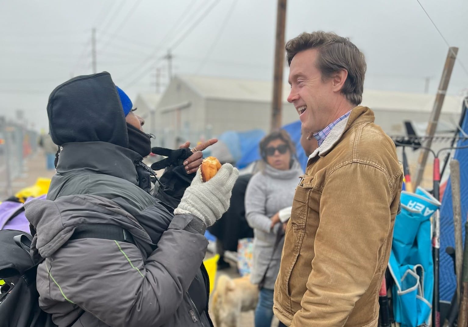 Denver, Colorado Mayor Mike Johnson smiles while speaking to a person in an outdoor location. They are both wearing winter clothing and the person talking to the mayor is holding a donut.