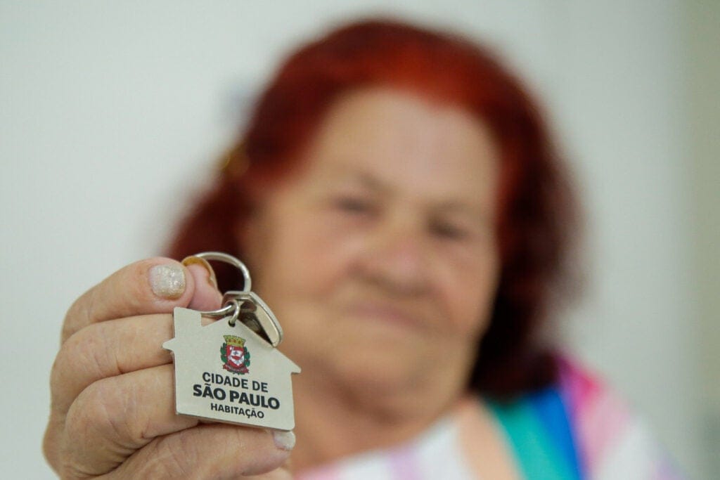 A soft focus, head and shoulders portrait of a woman in a colorful striped dress. In the foreground, her hand is in focus, holding a key ring with a metal, house-shaped keychain. The words on the keychain are in Portuguese and the translation is 'City of Sao Paulo Housing.'