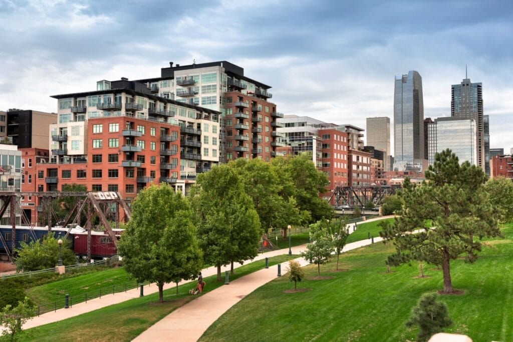The trees and grass of a Denver park are in the foreground. Apartment buildings, mostly red, occupy the middle of the frame, curving toward a distant view of tall office buildings downtown.