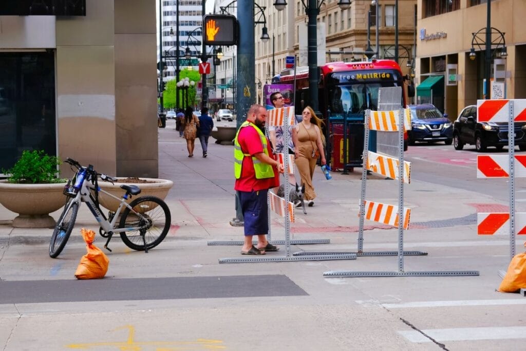 A busy street scene in downtown Denver includes a city worker in a fluorescent green vest, a pedestrian in a brown dress, a red city bus, and orange and white striped road barriers set up for an event.