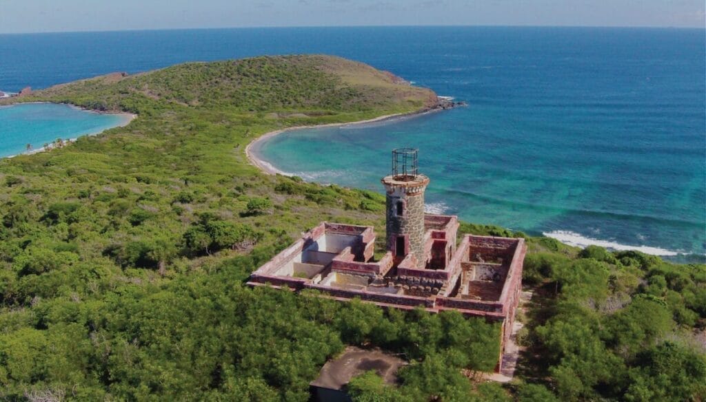 An aerial image of the Culebrita lighthouse in Puerto Rico, a brick structure with curving green coastland and blue ocean waters in the background,.