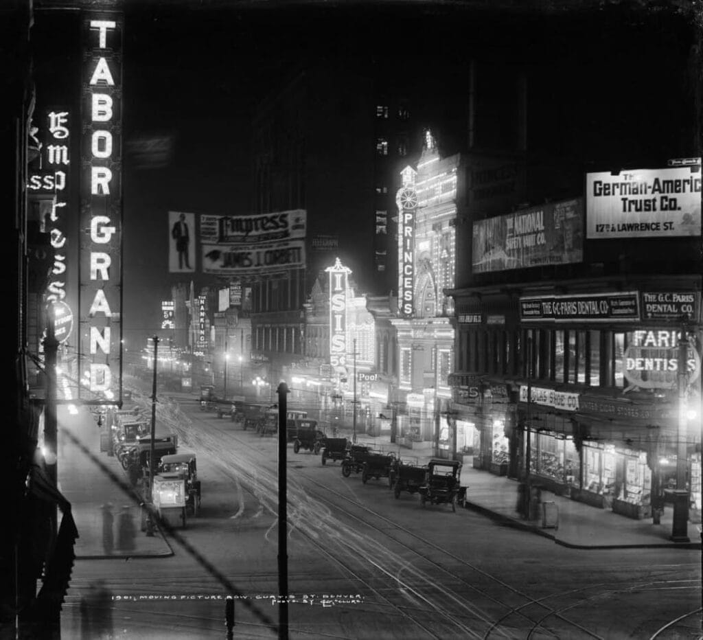 A black and white image of the Denver theater district in the early 1900s shows brightly lit theater marquees on both sides of a wide street, with automobiles of the period parked down the length of the street. Visible theater names include "Princess" and "Empress."