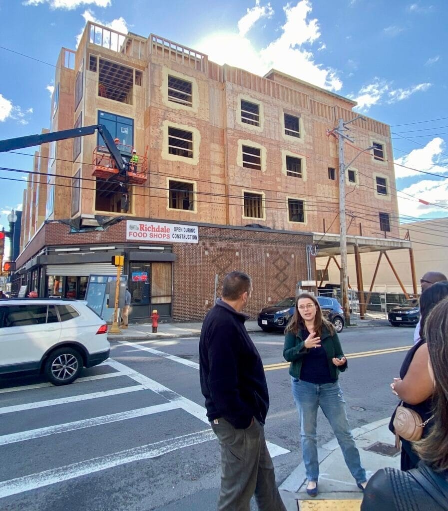 A woman in a dark jacket and jeans gestures as she speaks to a small, partially visible group on a street corner. Behind her a multi-story building is being constructed out of wood above a single-story brick food store. The sky is blue with a few scattered clouds.