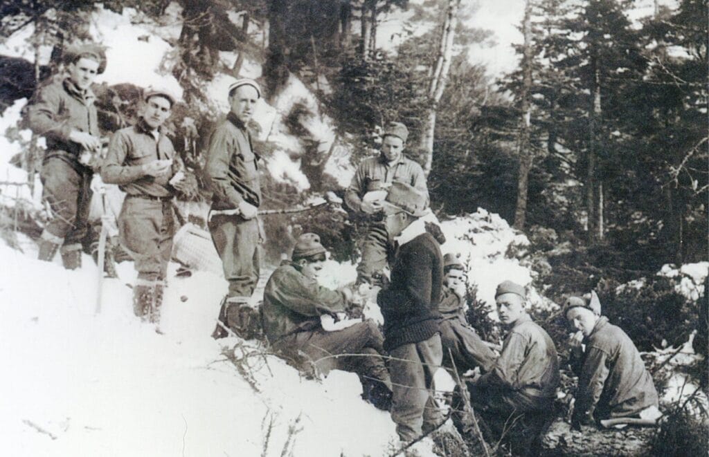 A black and white photo shows nine men from a Civilian Conservation Corps crew in the 1930s standing and sitting on a snowy hillside with trees in the background. They appear to be taking a break from their trailcutting work.