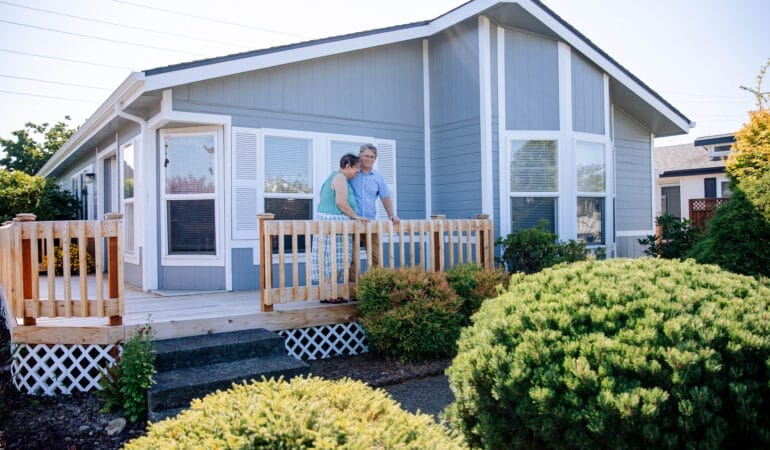 A man and woman stand on the porch of a manufactured home. The home is light blue with white trim and a peaked roof. Two large bushes stand in the foreground.