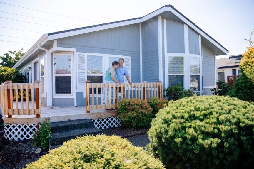 A man and woman stand on the porch of a manufactured home. The home is light blue with white trim and a peaked roof. Two large bushes stand in the foreground.