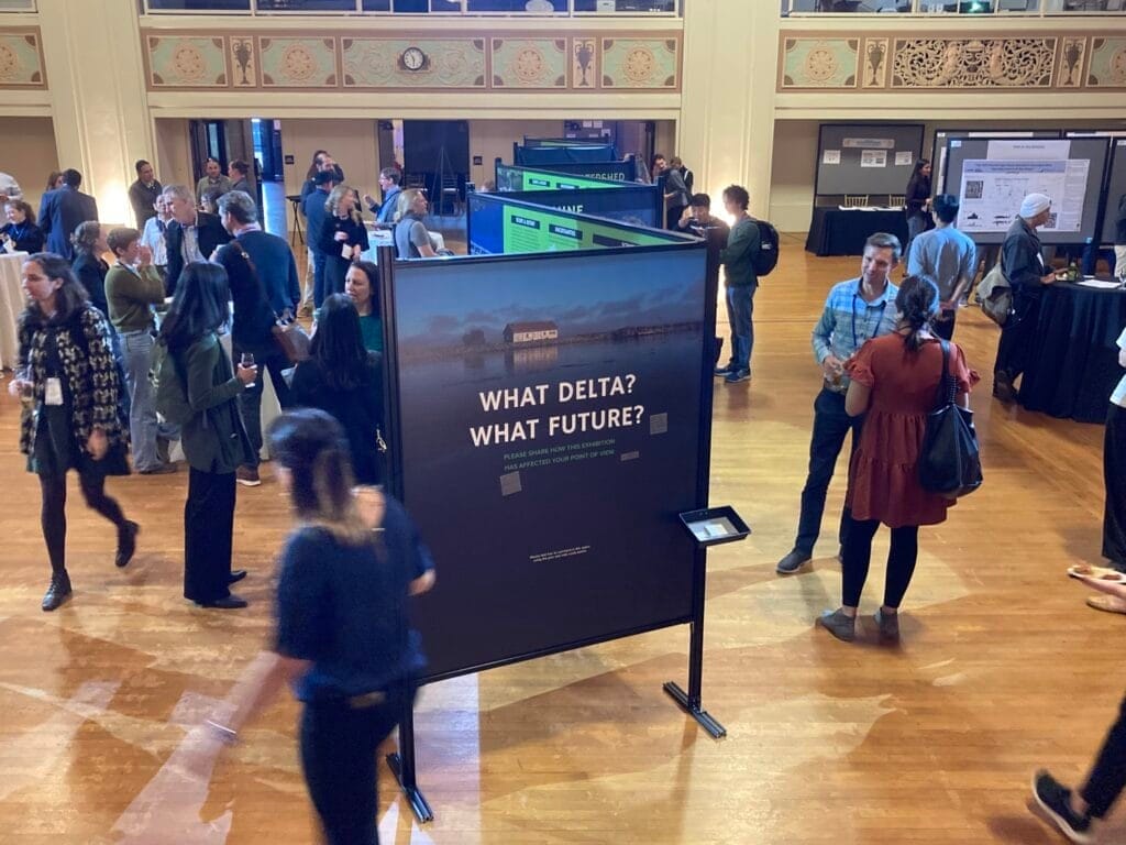 An aerial image of several people scattered around a large, wood-floored room, reading signs at a scenario planning workshop. The sign in the foreground reads, "What delta? What future?"