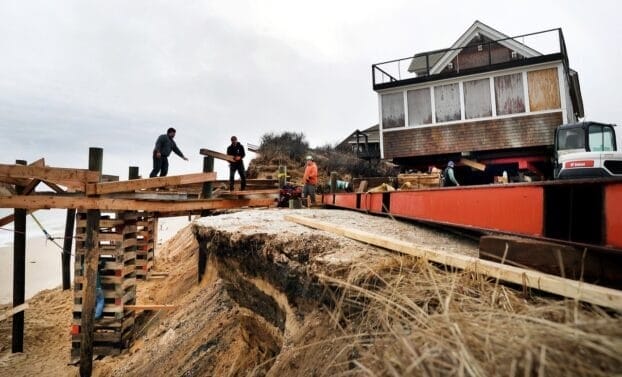 A photo of four men working outside of a house in Truro, Massachusetts. The men are putting the house on stilts to mitigate the encroaching threat of beach erosion.