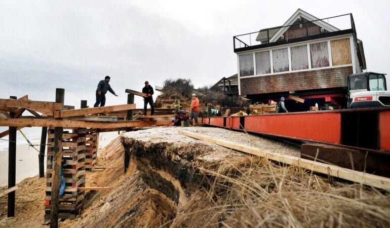 A photo of four men working outside of a house in Truro, Massachusetts. The men are putting the house on stilts to mitigate the encroaching threat of beach erosion.