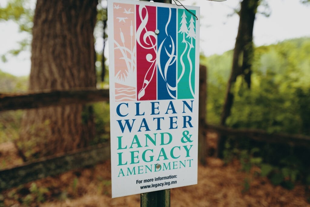 Colorful vertical rectangular icons on a Clean Water Land and Legacy Amendment sign. The icons are, from left to right, yellow with birds and cat-o-nine tails, red with a paintbrush and G clef, blue with a fish, and green with a trail running through two coniferous trees.